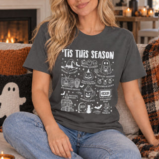 A woman sitting on a couch wearing a dark gray graphic t-shirt with white text reading 'TIS THIS SEASON' and various Halloween-themed illustrations like ghosts, pumpkins, a haunted house, and a cassette tape.