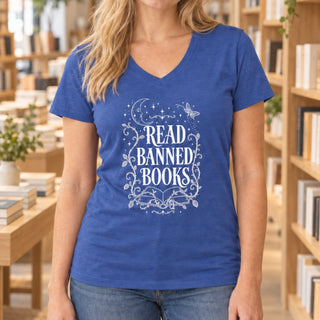 A woman wearing a heathered blue V-neck t-shirt with a white graphic that reads 'READ BANNED BOOKS' surrounded by decorative floral and celestial illustrations. The background is a softly blurred bookshop interior with wooden shelves and books.