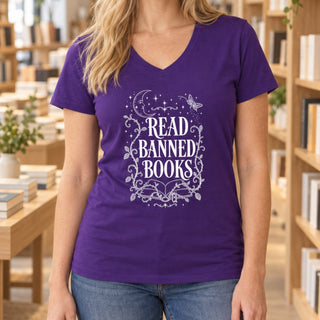 A woman wearing a purple V-neck t-shirt with a white graphic that reads 'READ BANNED BOOKS' surrounded by decorative stars, moons, and butterflies. The background shows a softly blurred library or bookstore setting with wooden bookshelves.