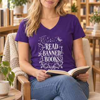 A woman sitting in a cozy armchair, wearing a purple V-neck t-shirt with a white graphic that reads 'READ BANNED BOOKS' surrounded by stars, moons, and butterflies. She is holding an open book in a warm, book-filled room.