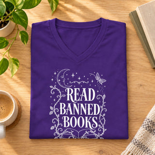 A folded purple V-neck t-shirt with a white graphic reading 'READ BANNED BOOKS' surrounded by stars, a crescent moon, and floral scrollwork, displayed on a wooden table with a plant, coffee cup, and books nearby.