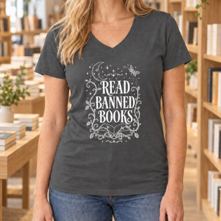 A woman wearing a dark gray v-neck t-shirt with a white graphic that reads 'READ BANNED BOOKS' surrounded by decorative stars, moons, and vines, set against a blurred library background.
