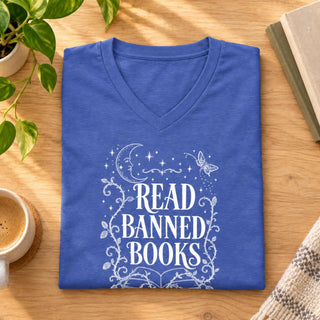 A folded blue V-neck t-shirt with a white graphic reading 'READ BANNED BOOKS' surrounded by stars, a crescent moon, and floral vines, displayed on a wooden table with a plant, coffee cup, and books nearby.