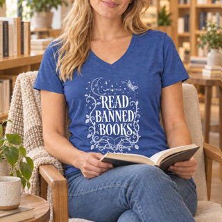 A woman sitting in a cozy armchair, holding an open book. She is wearing a heather blue v-neck t-shirt with a white graphic that reads 'READ BANNED BOOKS' surrounded by stars, moons, and butterflies.