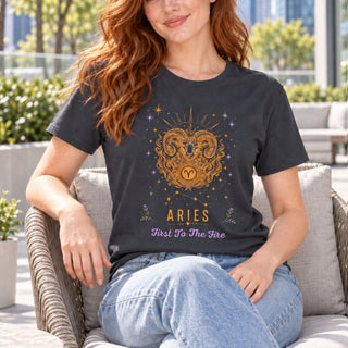 A woman sitting outdoors on a wicker chair wearing a dark charcoal gray graphic t-shirt featuring a golden ram design and the text 'ARIES First To The Fire'.