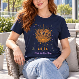 A woman sitting on a wicker patio chair wearing a navy blue graphic t-shirt with a gold Aries ram design and the text 'ARIES First To The Fire'.
