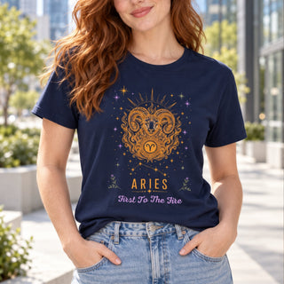A woman wearing a navy blue graphic t-shirt with a gold and purple Aries zodiac design, paired with light wash blue jeans, standing outdoors in a sunny, modern urban setting.