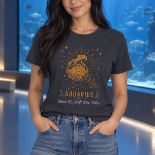 A woman wearing a dark charcoal gray graphic t-shirt with a gold and purple Aquarius design, paired with blue jeans, standing in front of a large blue aquarium window.