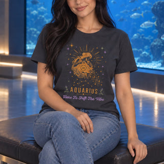 A woman wearing a dark charcoal gray graphic t-shirt with a gold and purple Aquarius design, sitting on a black leather bench in front of a large aquarium.