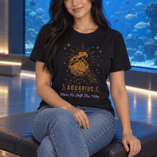 A woman wearing a black graphic t-shirt with a gold and purple Aquarius zodiac design, sitting on a black leather bench in front of a large blue aquarium.