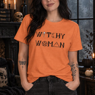 A woman wearing a bright orange t-shirt with a black graphic that reads 'WITCHY WOMAN' in a mystical font, set against a dark, gothic-themed background with candles, skulls, and pumpkins.