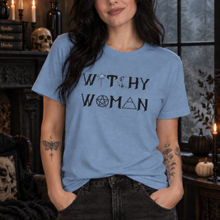 A woman wearing a light blue graphic t-shirt with the words 'WITCHY WOMAN' printed in black gothic-style lettering, posed in a dark, atmospheric room with candles and Halloween decor.