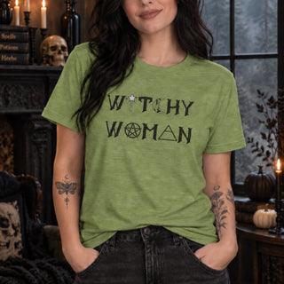 A woman wearing a heathered sage green t-shirt with a black graphic that reads 'WITCHY WOMAN' with occult symbols, standing in a dark, atmospheric room with candles and Halloween decorations.