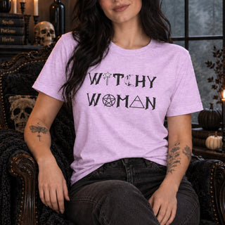 A woman wearing a light purple heather t-shirt with the words 'WITCHY WOMAN' printed in black gothic-style lettering with occult symbols, sitting in a dark, moody room with Halloween decorations.
