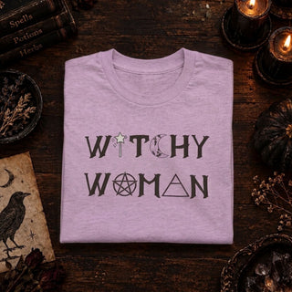 A folded light lavender t-shirt with a gothic 'WITCHY WOMAN' graphic print, displayed on a dark rustic wooden table surrounded by witch-themed props like candles, a black bird illustration, and old books.