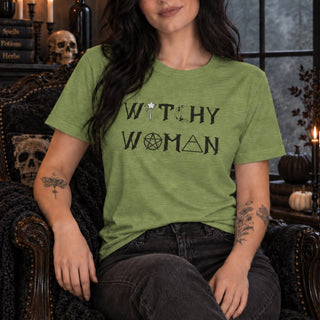 A woman wearing a sage green graphic t-shirt with the words 'WITCHY WOMAN' printed in black, sitting in a dark, gothic-style armchair with skulls and candles in the background.