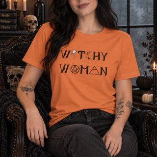 A woman wearing a bright orange graphic t-shirt with the words 'WITCHY WOMAN' printed in black with occult symbols, sitting in a dark, gothic-style armchair.