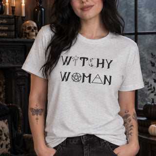 A woman wearing a light gray heather t-shirt with the text 'WITCHY WOMAN' printed in black with occult-themed symbols, set against a dark, gothic room background.