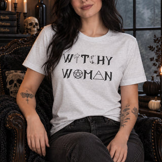 A woman wearing a light gray heather t-shirt with a black graphic print that reads 'WITCHY WOMAN' using occult symbols, seated in a dark, gothic-themed armchair.