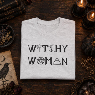 A folded light gray heather t-shirt with a black graphic reading 'WITCHY WOMAN' using occult symbols, displayed on a dark rustic wooden table surrounded by candles, a small pumpkin, books, and dried flowers.