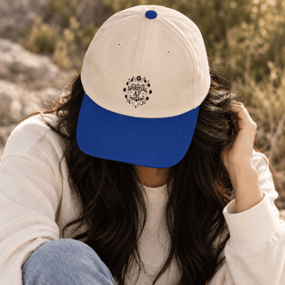 A person outdoors wearing a baseball cap with an off-white crown and a royal blue brim. The cap has a small circular black graphic on the front and a blue button on top.