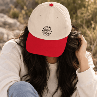 A person outdoors wearing a cream and red baseball cap with a black circular graphic on the front. The cap has a red visor, red button on top, and the person is wearing a cream sweatshirt and blue jeans.