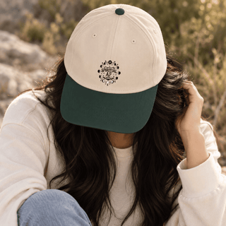 A person outdoors wearing a two-tone baseball cap with an off-white crown and a dark green brim. The front of the cap features a circular black graphic with the text 'BORN TO BE'.