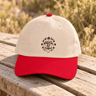 A baseball cap with an off-white crown and a bright red curved brim, resting on a weathered wooden bench with a blurred outdoor background. The front of the cap features a black circular graphic with skulls, roses, and the text 'SPIRITUAL AF'.