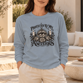 A woman wearing a light blue long-sleeve t-shirt with a black graphic print of an all-seeing eye, roses, candles, and the text 'PROTECTED BY MY ANCESTORS'. She is standing in a sunlit outdoor patio setting.