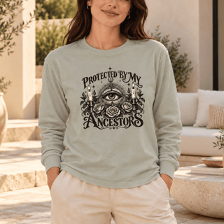 A woman wearing a sage green long-sleeve t-shirt with a black graphic print that reads 'PROTECTED BY MY ANCESTORS' around an all-seeing eye and roses, set against a warm, sunlit patio background.