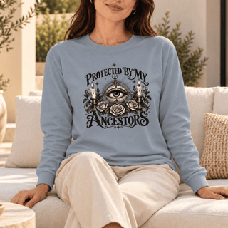 A woman wearing a light blue long-sleeve graphic sweatshirt sits on a white sofa in a bright, modern living room. The sweatshirt features a black graphic of an all-seeing eye surrounded by roses and candles with the text 'PROTECTED BY MY ANCESTORS'.