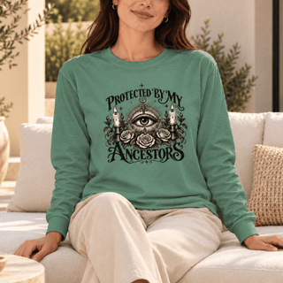 A woman wearing a sage green long-sleeve t-shirt with a black graphic print that reads 'PROTECTED BY MY ANCESTORS' around an all-seeing eye, candles, and roses, sitting on a white outdoor sofa.