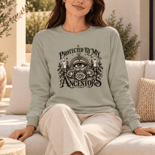 A woman sits on a light-colored outdoor sofa wearing a sage green long-sleeve crewneck sweatshirt with a black graphic print that reads 'PROTECTED BY MY ANCESTORS 2025' around an all-seeing eye and roses. She is wearing cream-colored pants.
