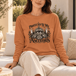 A woman sitting on a light-colored outdoor sofa wearing a rust-orange crewneck sweatshirt with a black graphic print that reads 'PROTECTED BY MY ANCESTORS 1977' around an all-seeing eye and roses design.