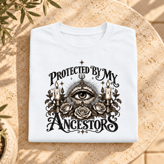A folded white t-shirt with a black gothic-style graphic reading 'PROTECTED BY MY ANCESTORS' around an all-seeing eye, candles, and roses, displayed on a round stone table with leaf shadows.