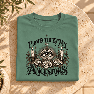A folded sage green t-shirt with a detailed graphic print reading 'PROTECTED BY MY ANCESTORS' around an all-seeing eye, roses, and candles, displayed on a round stone table.