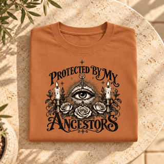 A folded terracotta orange t-shirt with a black gothic graphic reading 'PROTECTED BY MY ANCESTORS' around an all-seeing eye, candles, roses, and stars, displayed on a round stone table with soft plant shadows.