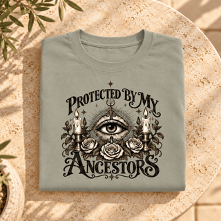 A folded sage green t-shirt with a detailed black graphic print reading 'PROTECTED BY MY ANCESTORS' around an all-seeing eye, candles, and roses, displayed on a textured stone surface with soft plant shadows.