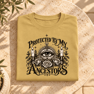 A folded tan t-shirt with a black graphic print reading 'PROTECTED BY MY ANCESTORS' around an all-seeing eye, candles, and roses, displayed on a round stone table.