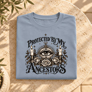 A folded light blue t-shirt with a gothic-style graphic reading 'PROTECTED BY MY ANCESTORS' around an all-seeing eye, candles, and roses, displayed on a round stone table.