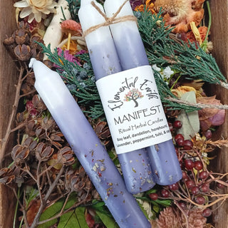 Two lavender herbal candles tied together with twine and one matching taper candle displayed on a bed of dried flowers, greenery, and berries. The label reads 'Flower Fairy Manifest Ritual Herbal Candles'.