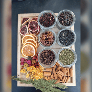 An overhead flat-lay of a wooden tray filled with assorted dried botanicals and spices in small clear jars, including citrus slices, star anise, cinnamon sticks, dried flowers, herbs, and seeds, set on a dark surface.