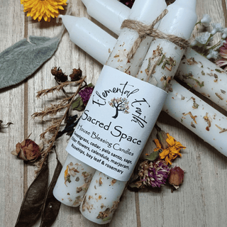 Three white wax candles tied with twine and wrapped in a white paper label reading 'Elemental Fire Sacred Space House Blessing Candles,' arranged on a light wood surface with dried flowers and botanicals.