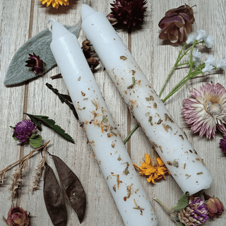 Two white taper candles with dried botanical inclusions, arranged on a light wood surface surrounded by decorative dried flowers and leaves.