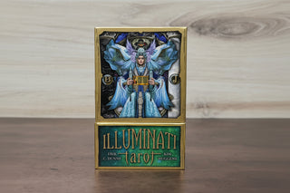 A boxed tarot deck standing upright on a wooden surface against a light wood wall. The box has a gold border and a front panel showing a blue-toned illustrated figure with wings, and the lower section reads 'ILLUMINATI tarot' with creator names below.