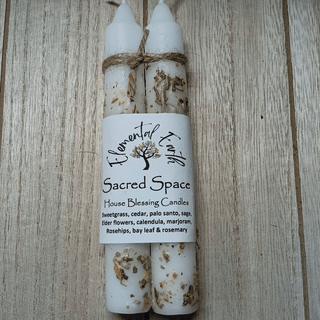 Two white beeswax candles bundled together with a twine bow and a white paper label that reads 'Flowerful Trees Sacred Space House Blessing Candles,' placed on a light wood surface.