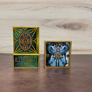 Two small decorative card boxes or cards displayed on a wooden surface against a light wood wall. The left item is labeled 'ILLUMINATI tarot' and shows a gold-toned geometric design with teal and green accents.