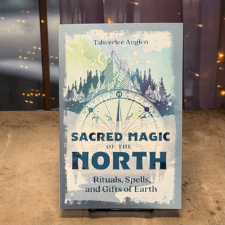 A book titled "Sacred Magic of the North" by Tahverlee Anglen, displayed upright on a small stand. The cover features a mystical illustration with a compass, mountains, stars, and a crescent moon in soft blue, white, and gray tones.