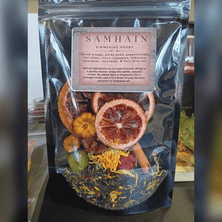 A black resealable pouch of Samhain Simmering Herbs tea blend, showing dried citrus slices, herbs, and spices through a clear window, with a paper label on the front.