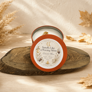 A round candle tin with its lid off, placed on a rustic wood slice. The lid label reads 'Smells Like a Waxing Moon' and 'Harvest Moon.